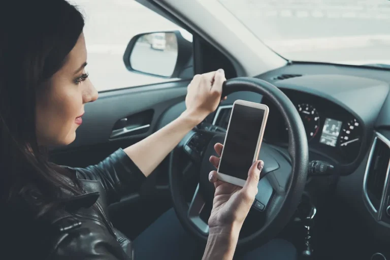 Young woman looking to her smartphone while driving car at front window background. Distracted female driver. Concept of dangerous driving. Woman driving and holding cellphone with blank screen.