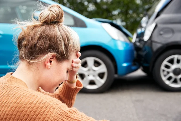 Female Motorist With Head In Hands Sitting Next To Vehicles Involved In Car Accident